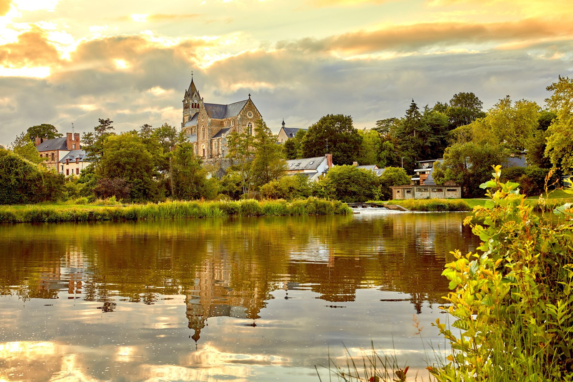 Une rivière calme reflète une église et des arbres sous un ciel nuageux et doré.