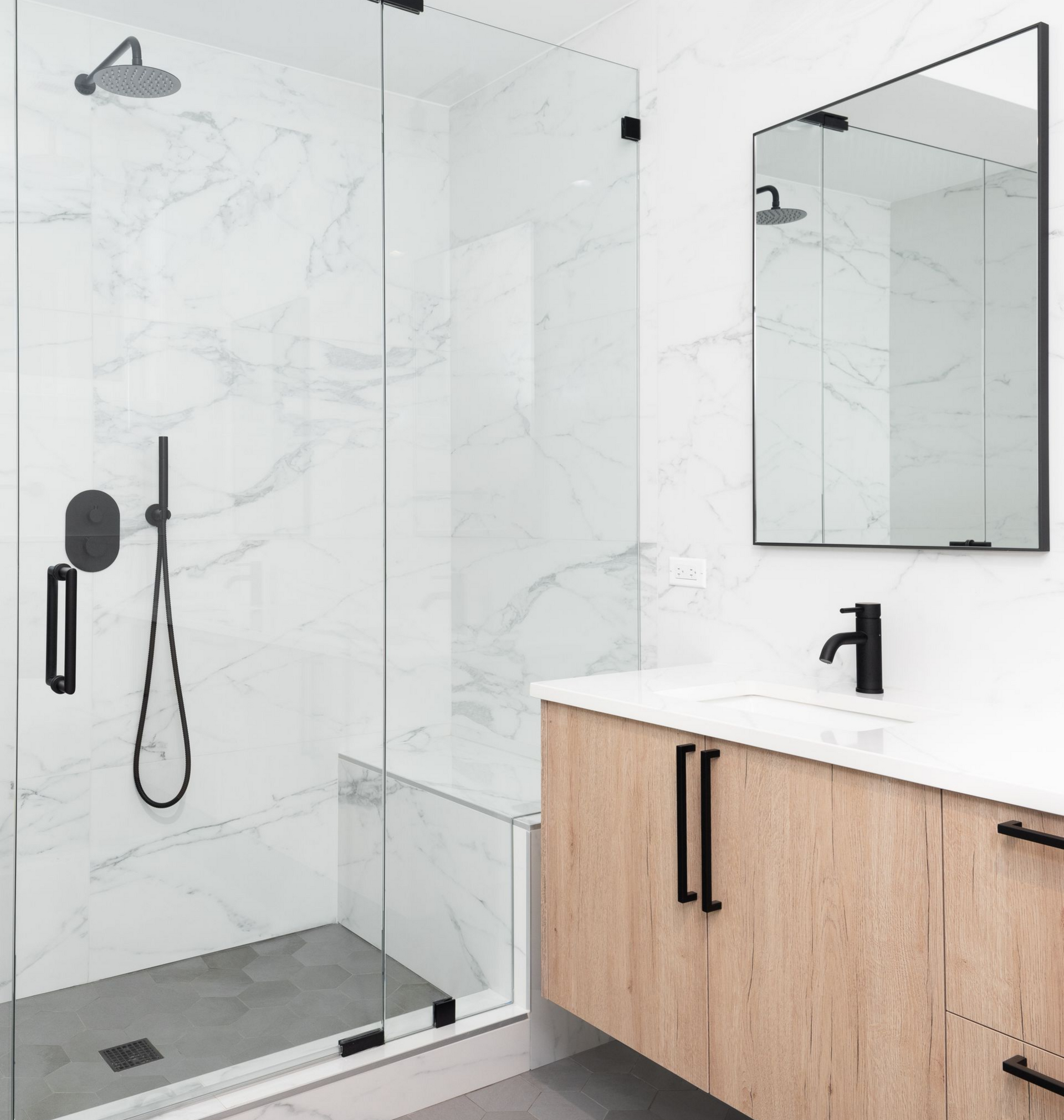 Salle de bains moderne avec douche carrelée en marbre, cabine en verre, vanité en bois clair, luminaires noirs et miroir rectangulaire.