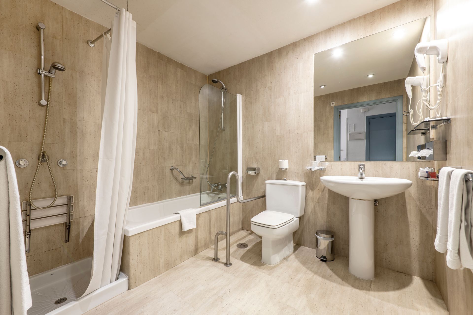 Salle de bains avec baignoire, douche, toilettes et lavabo. Tons neutres, avec barres d'appui accessibles.