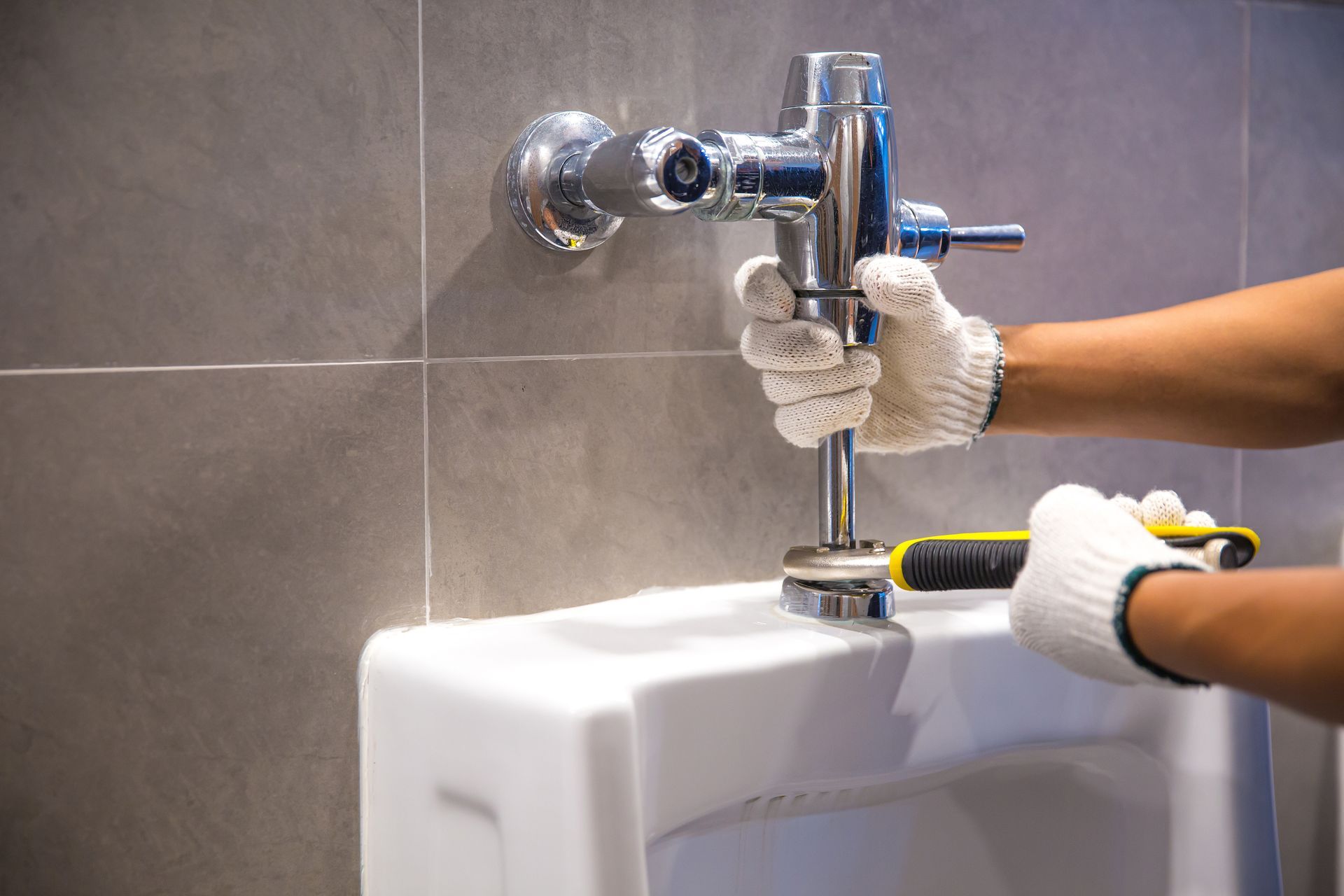 Un plombier installe un robinet de chasse chromé sur un urinoir blanc dans une salle de bains carrelée.