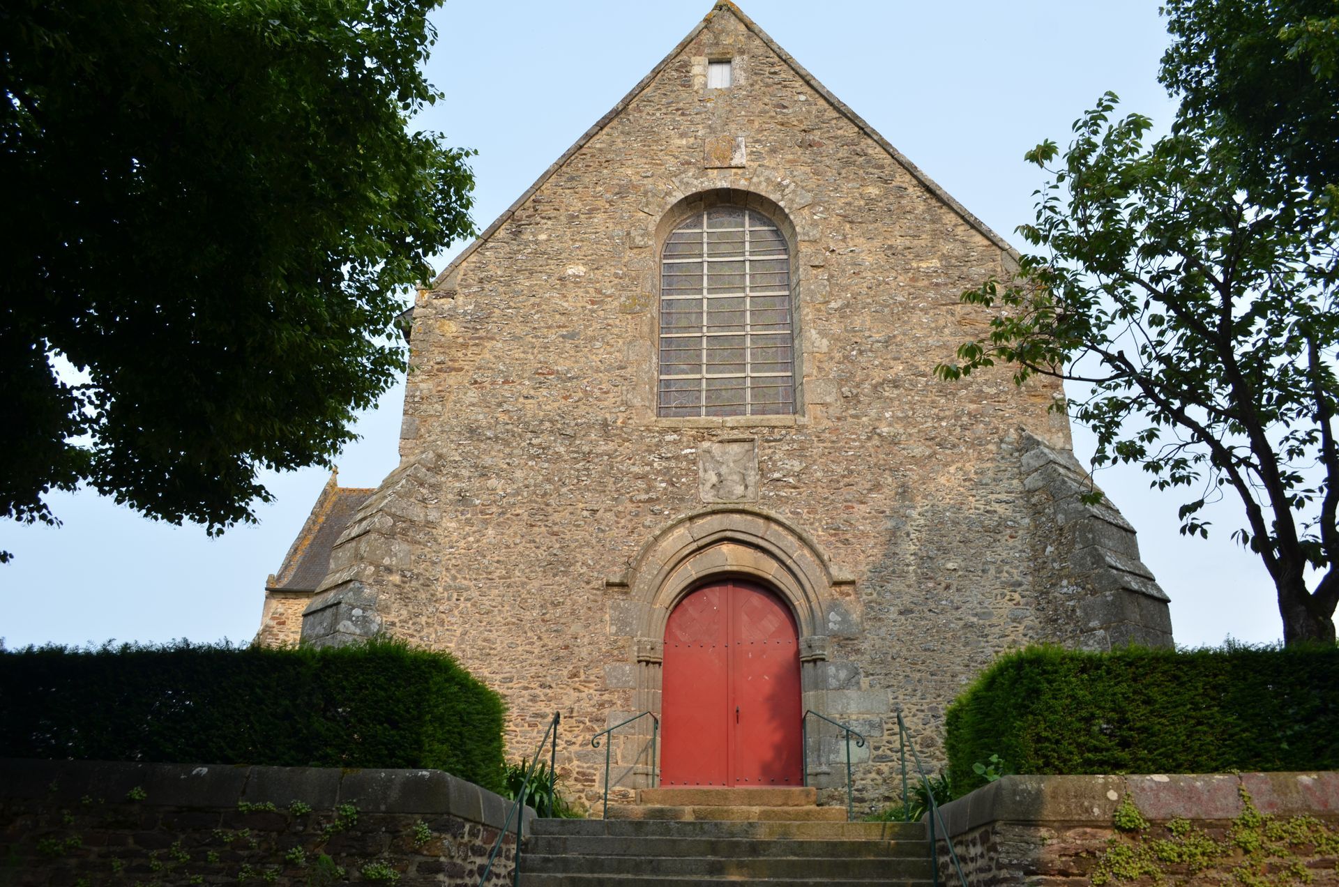 Façade d'église en pierre avec porte rouge, fenêtre cintrée et arbres.