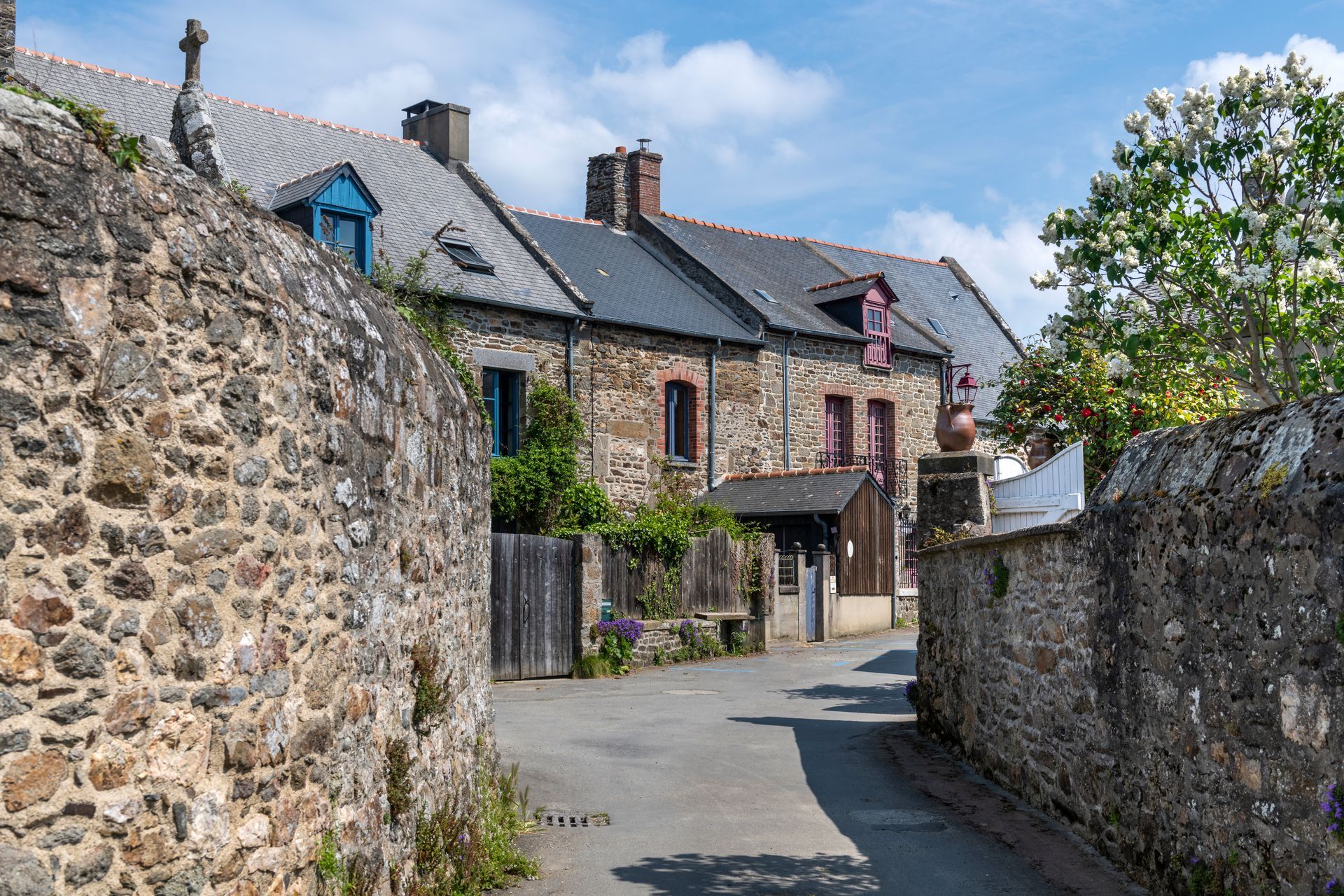 Des bâtiments en pierre bordent une rue étroite sous un ciel bleu ; une porte est visible entre les murs de pierre.