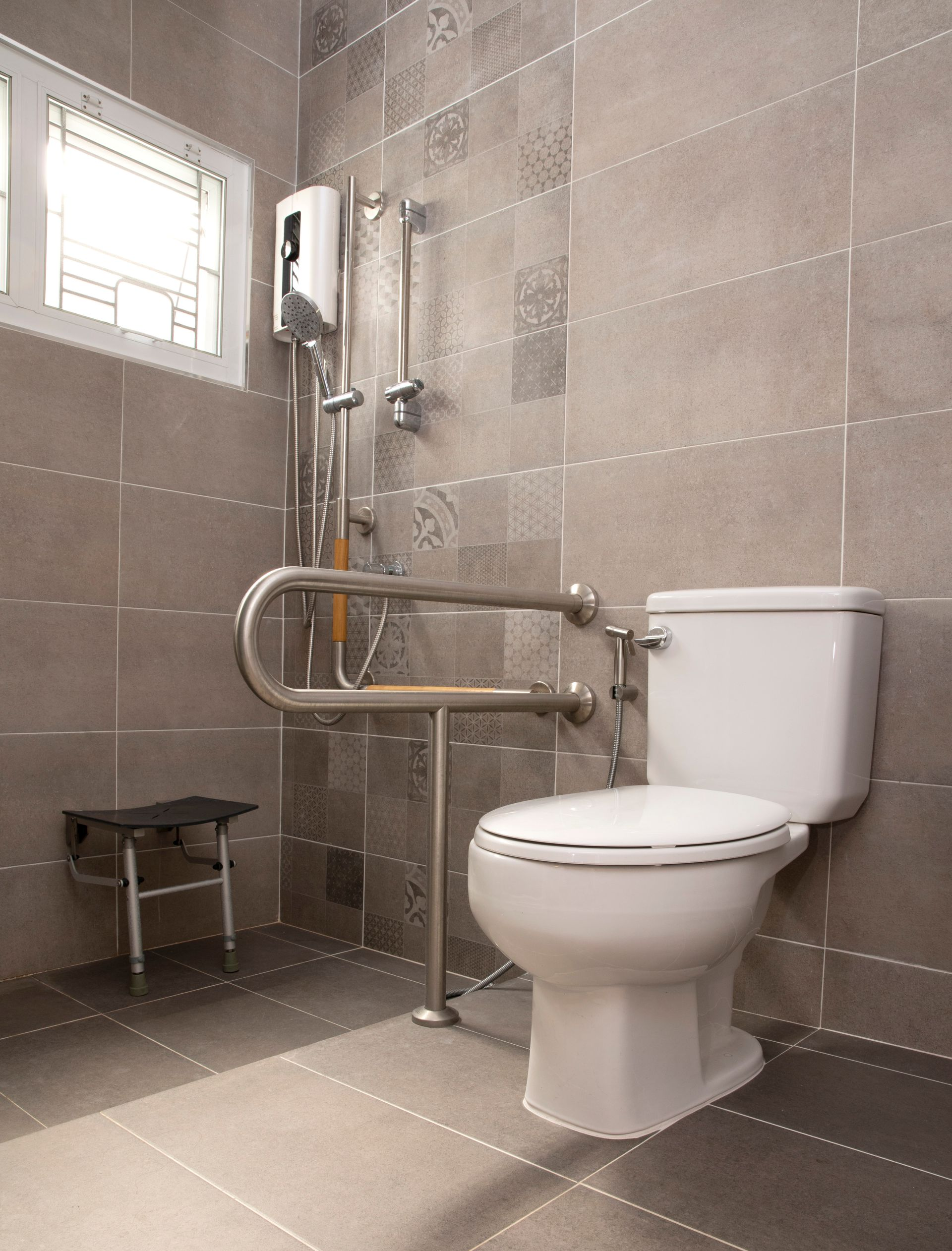 Salle de bains avec w.-c., barre d'appui, douche, petit tabouret et carrelage gris.