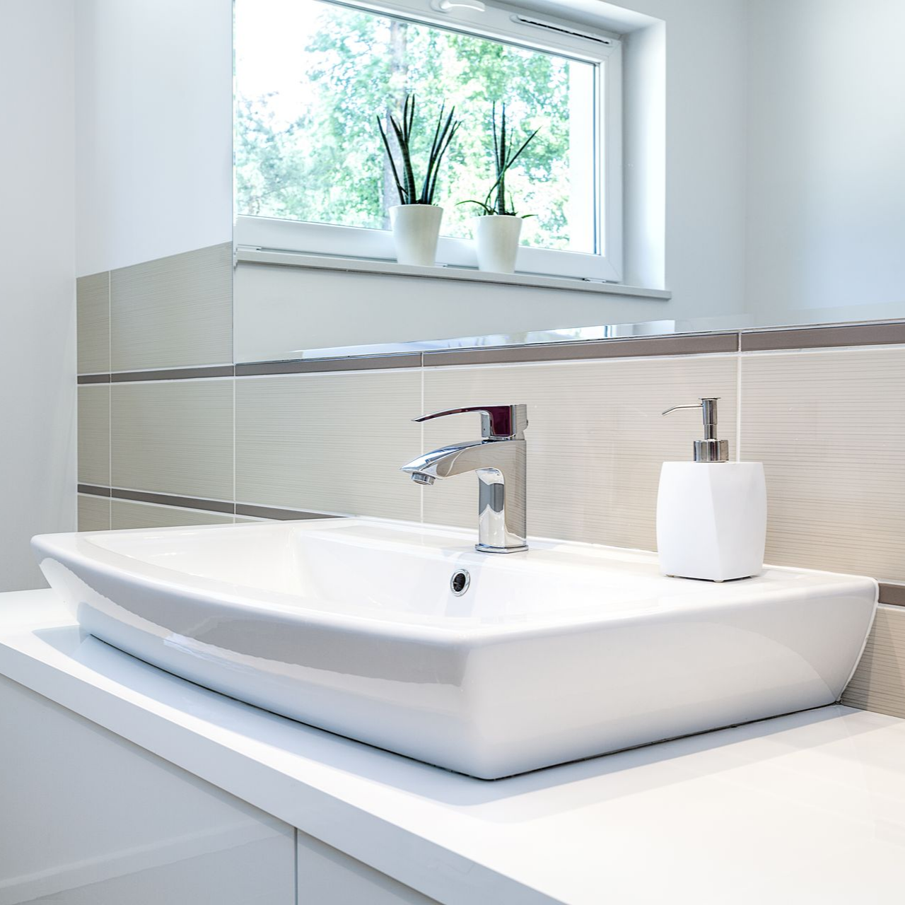 Lavabo blanc avec robinet chromé et distributeur de savon. Placé près d'une fenêtre ornée de plantes vertes.