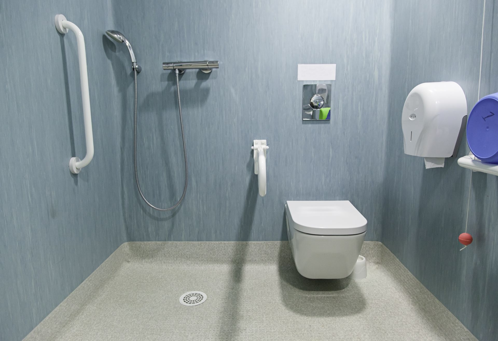 Salle de bains accessible avec douche, toilettes, barres d'appui et distributeur sur des murs texturés bleus.