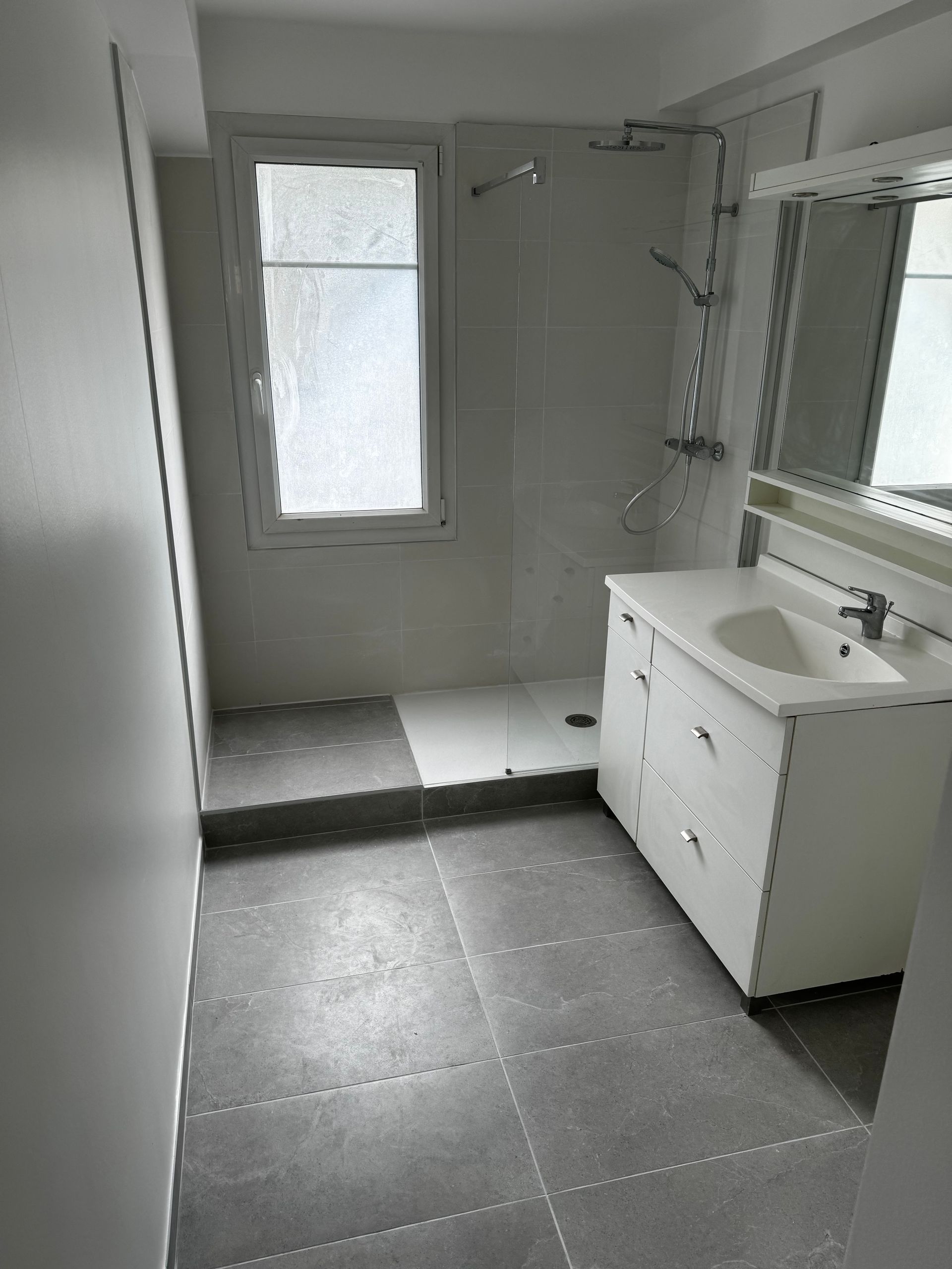 Salle de bains avec sol carrelé gris, vanité blanche, douche et fenêtre.