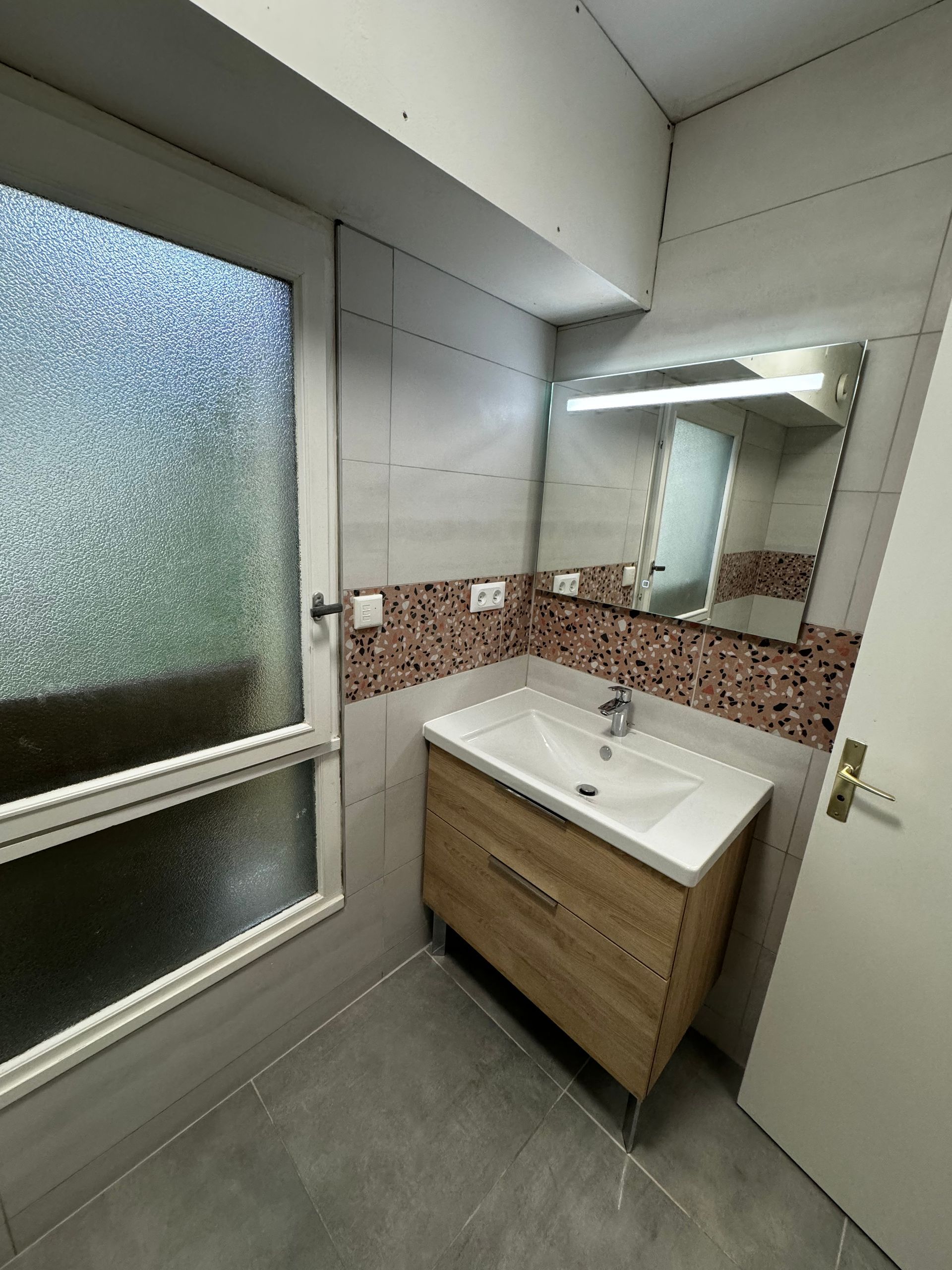 Salle de bains avec une vanité en bois, un grand miroir et une fenêtre givrée.