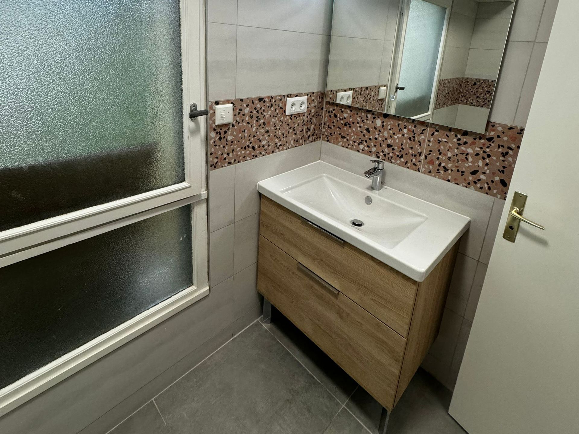 Salle de bains avec meuble lavabo en bois, lavabo blanc, miroir et mur texturé. Fenêtre à gauche.