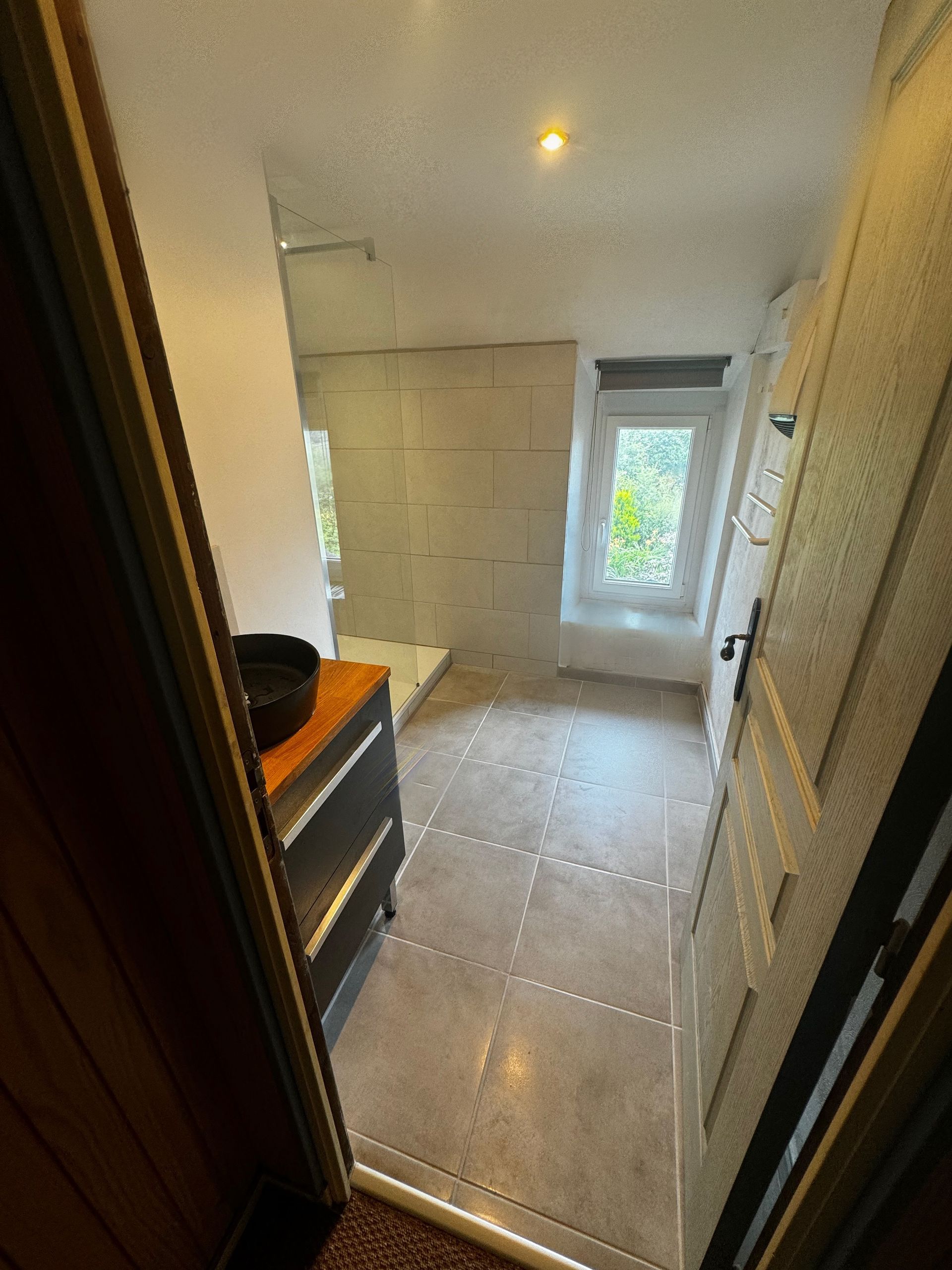 Salle de bains avec fenêtre, douche et meuble noir. Carrelage et murs gris clair, porte blanche.