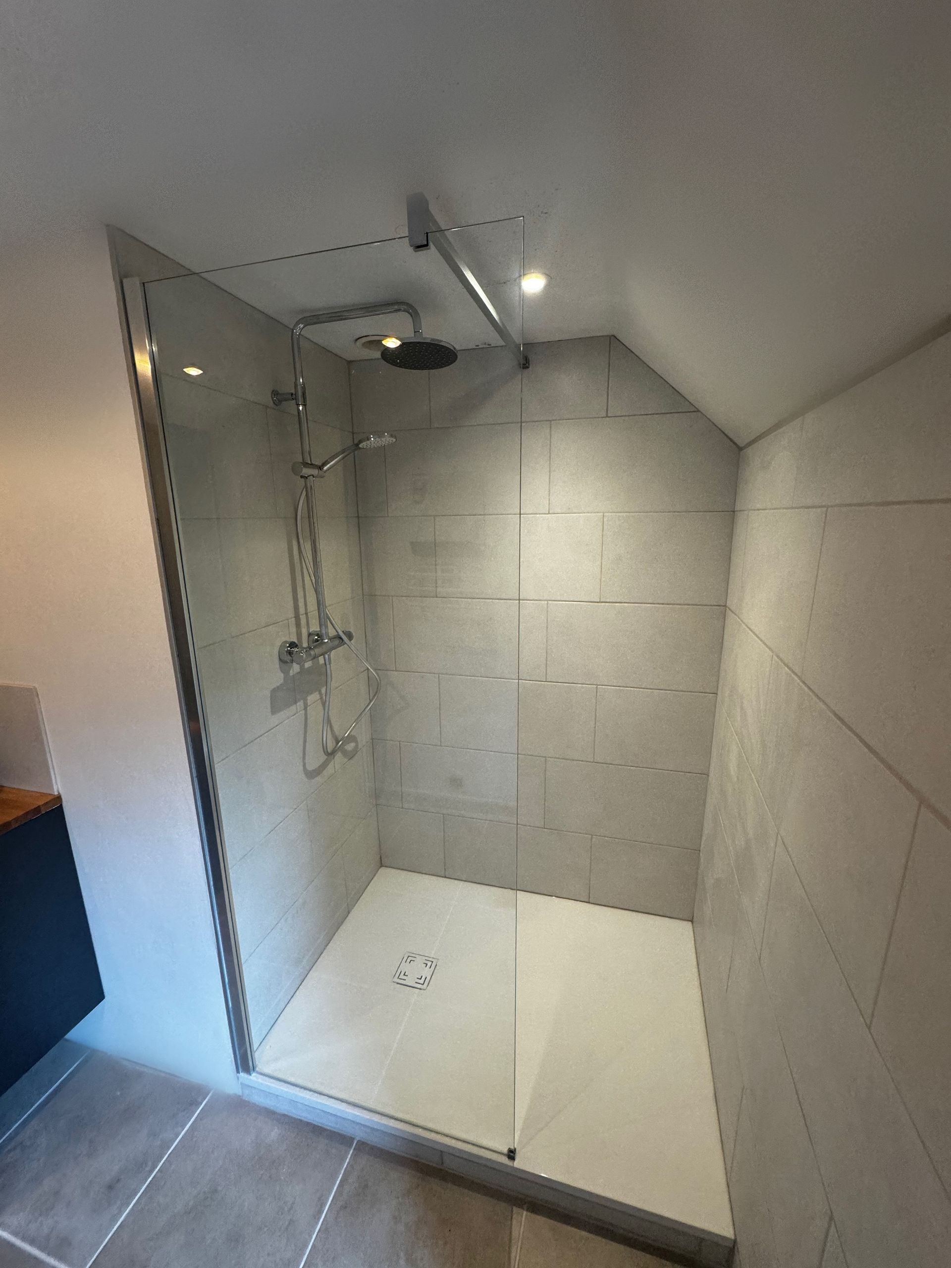 Cabine de douche moderne avec porte vitrée, murs carrelés gris et base de douche blanche.
