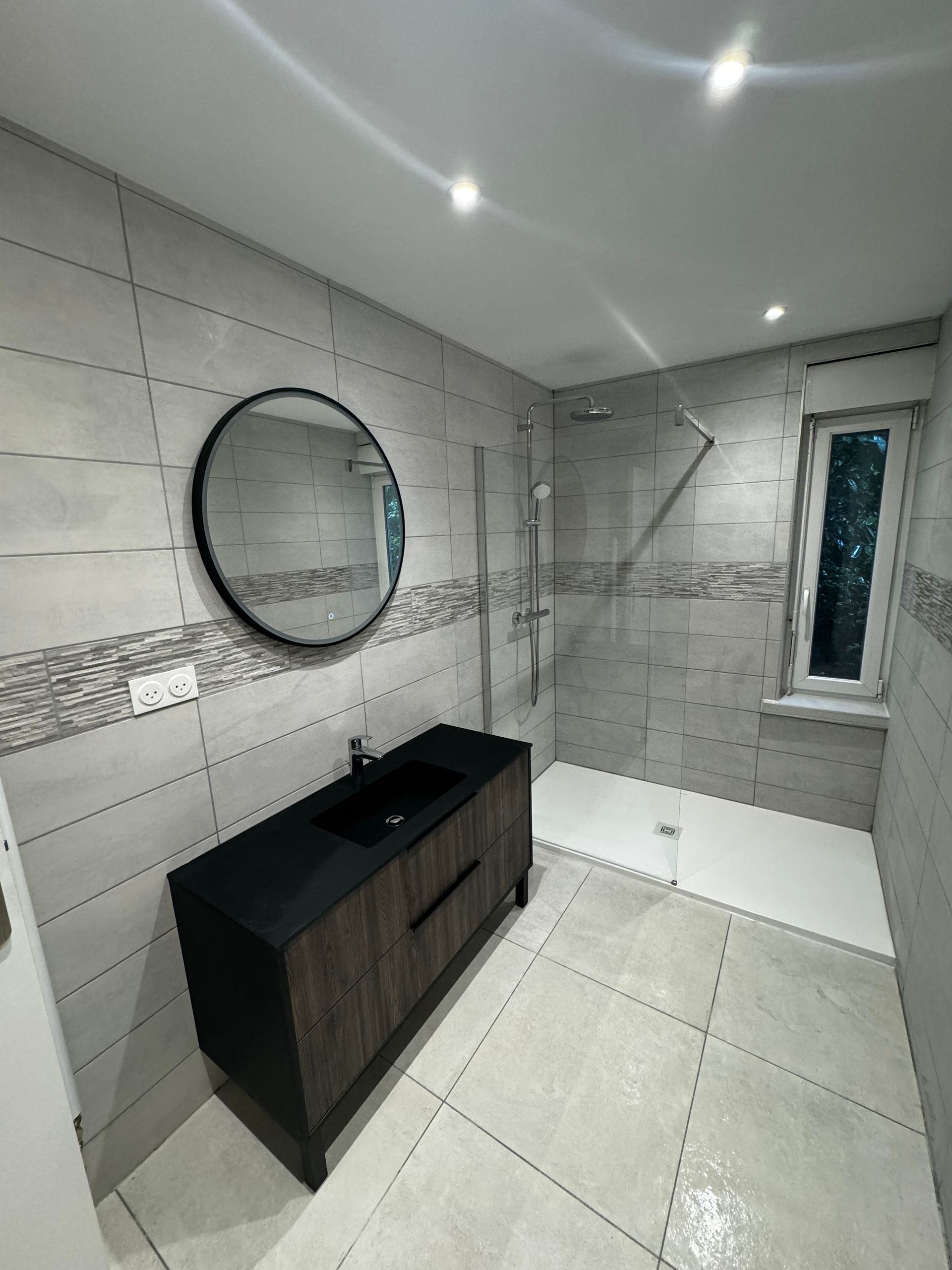 Salle de bains moderne : meuble lavabo avec dessus noir, miroir rond, douche, carrelage gris, petite fenêtre.