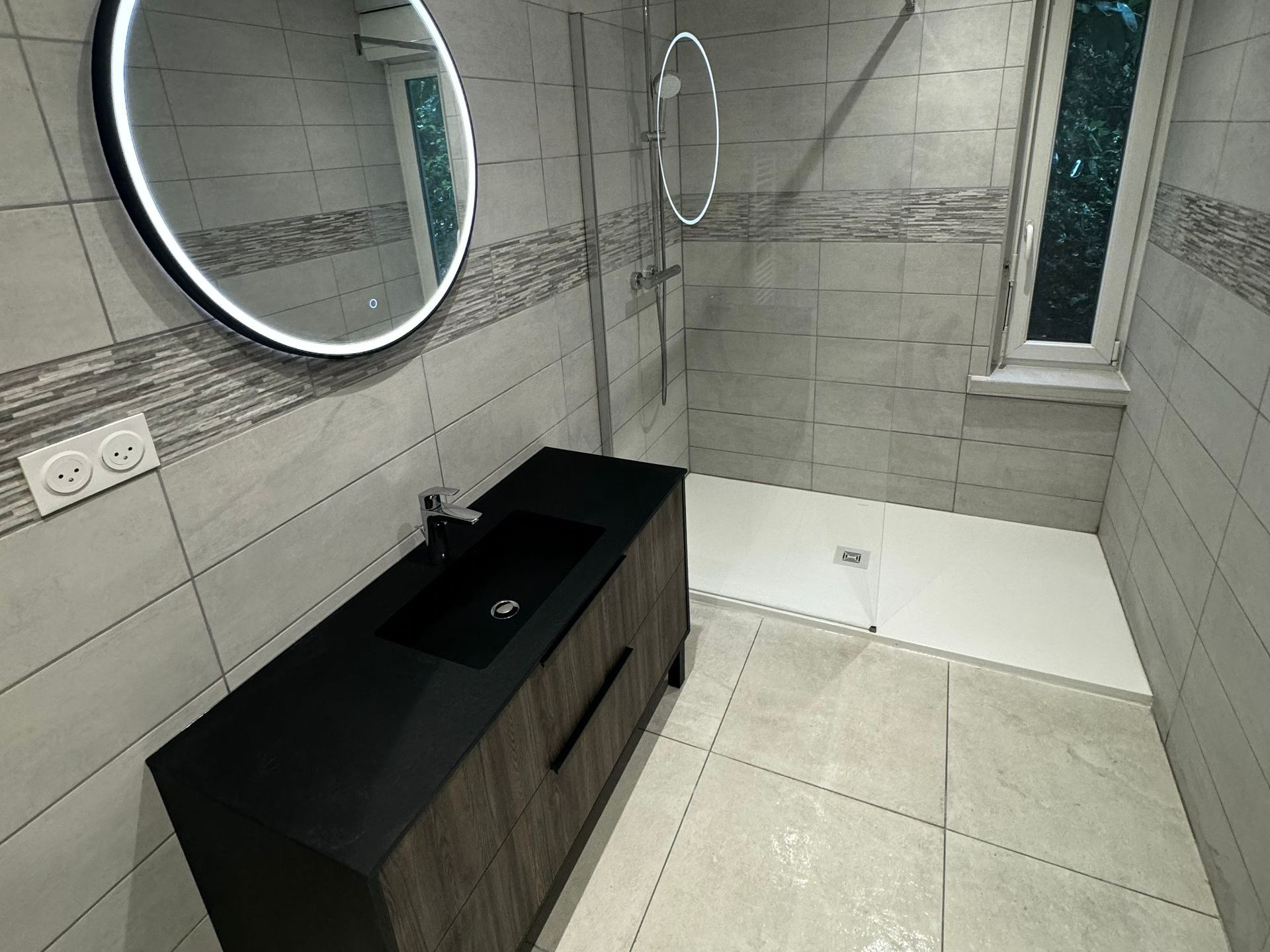 Salle de bains moderne avec une vanité sombre, une douche, un miroir rond et une fenêtre.