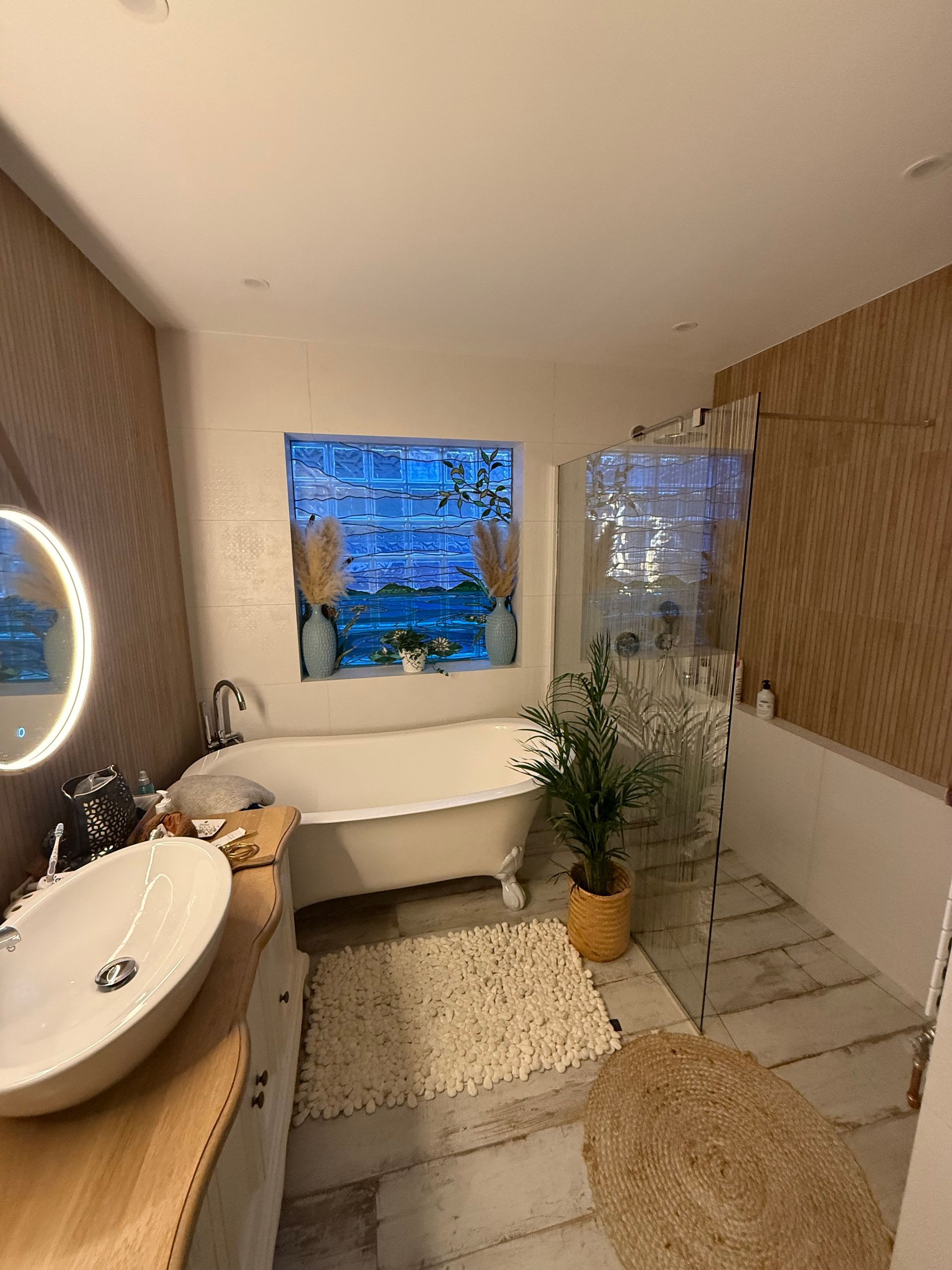Salle de bains avec baignoire sur pieds, grande fenêtre et meuble lavabo en bois clair. Tons neutres, tapis et plante en pot.