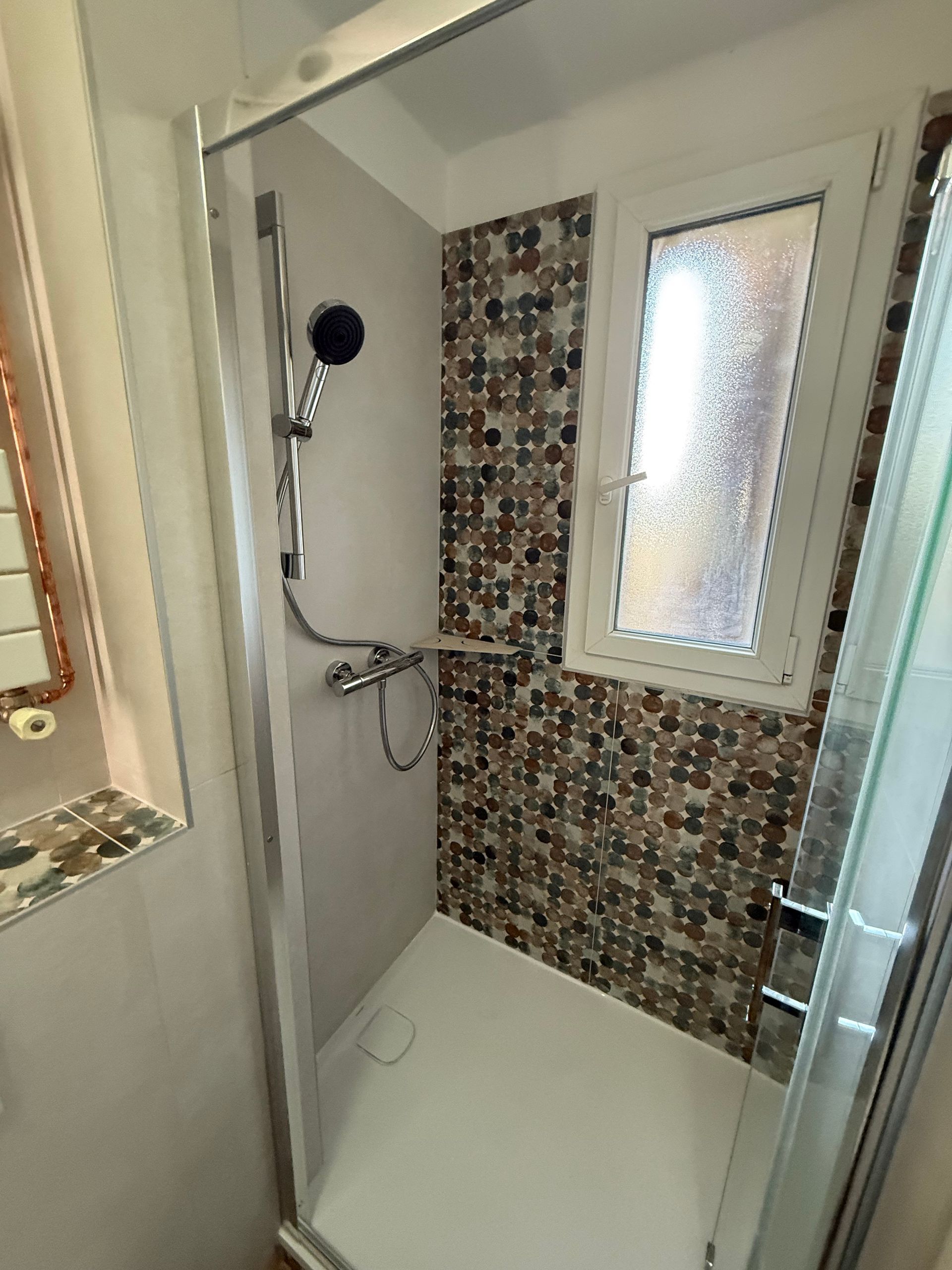 Salle de bains moderne avec douche en verre, mur en carrelage gris et toilettes blanches.