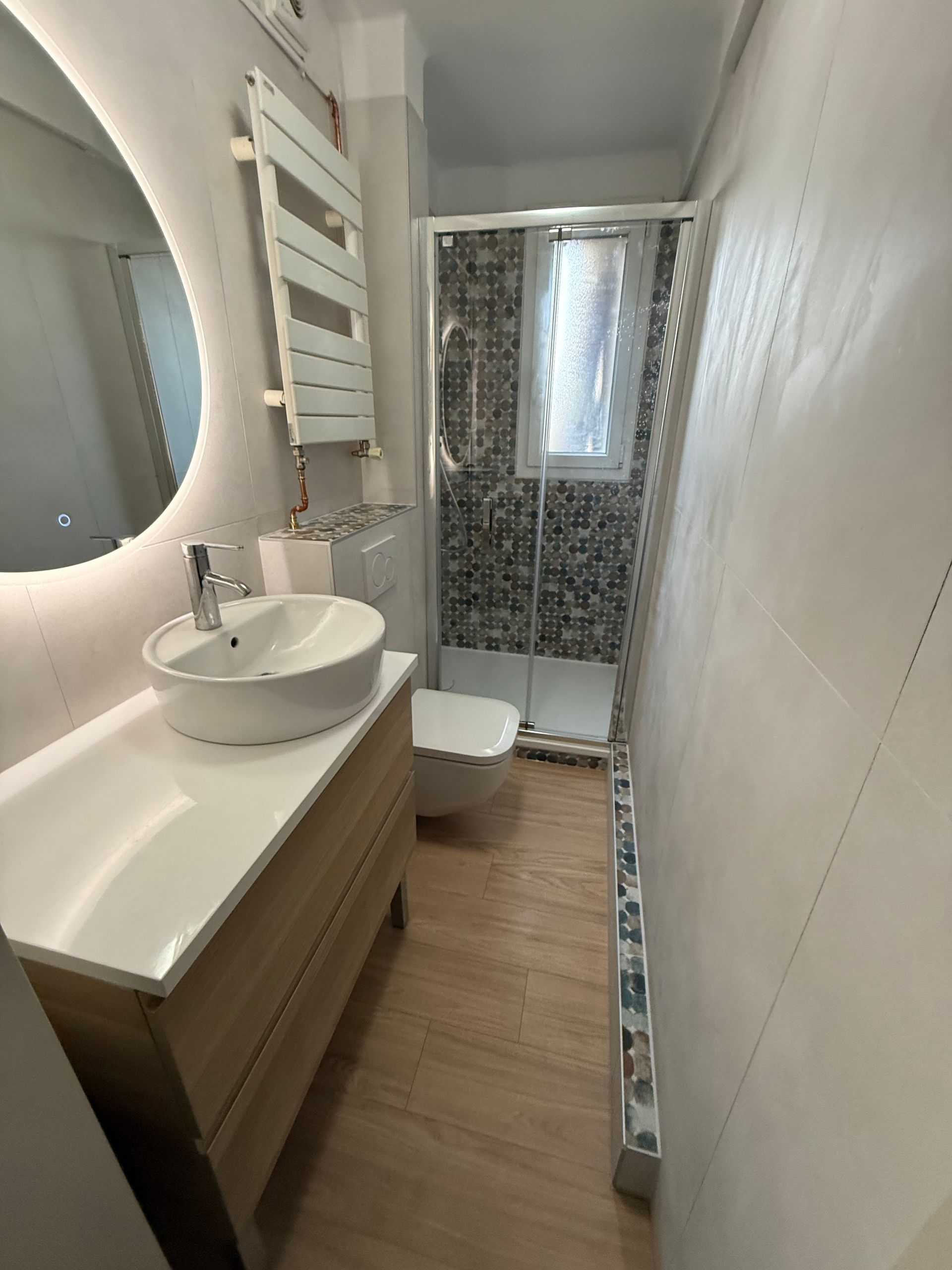 Salle de bains moderne avec douche en verre, mur en carrelage gris et toilettes blanches.