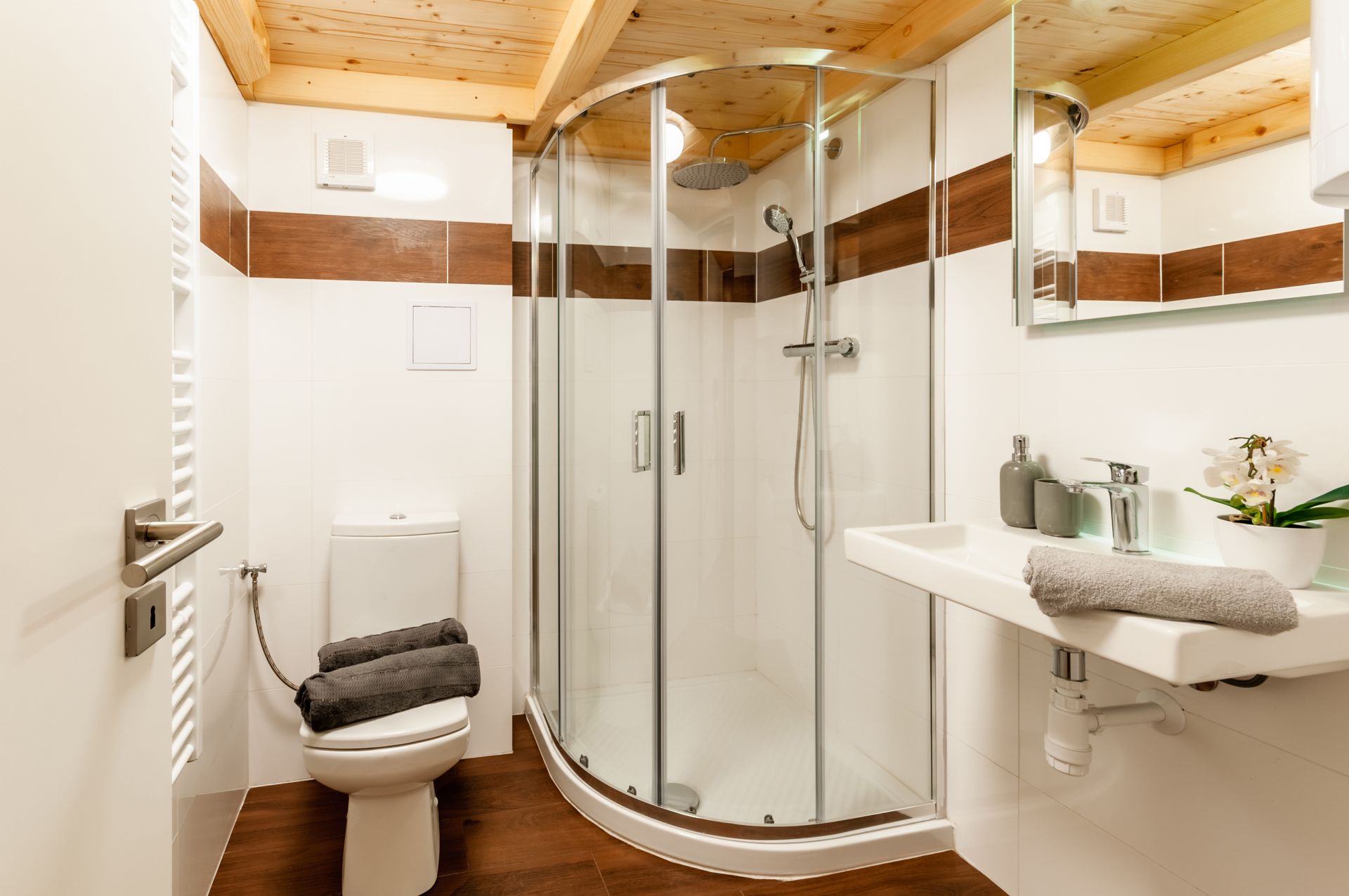 Salle de bains avec douche en verre incurvé, toilettes, lavabo et plafond en bois. Couleurs marron et blanc.