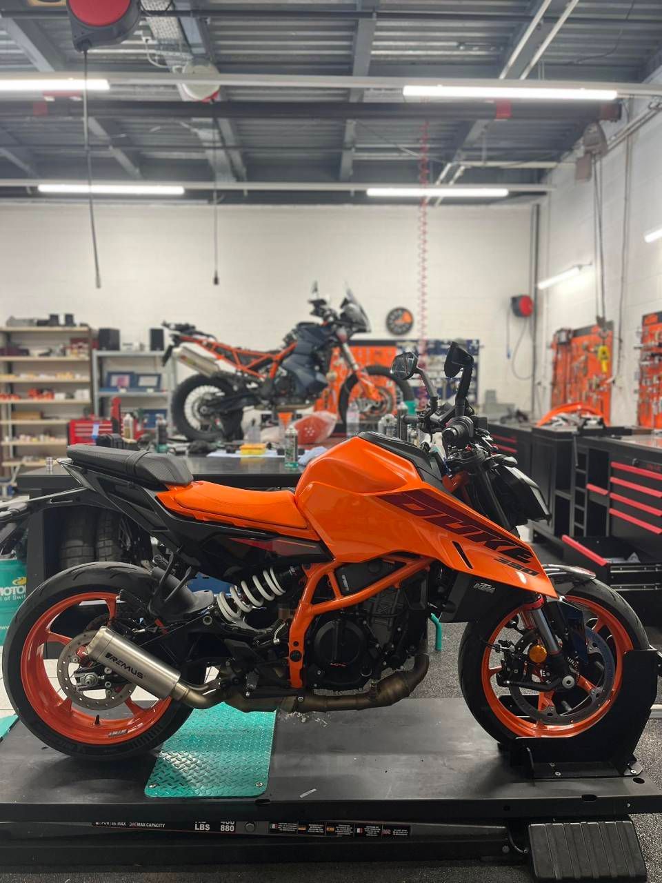 Une moto KTM orange et noire dans un atelier, une autre moto en arrière-plan.