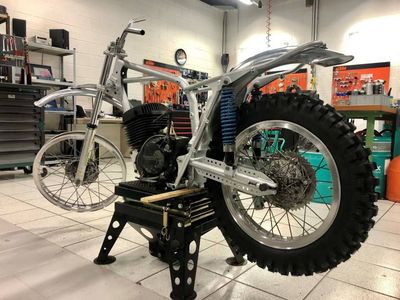 Moto cross vintage partiellement assemblée sur un support noir, cadre argenté, moteur noir, gros pneu arrière.