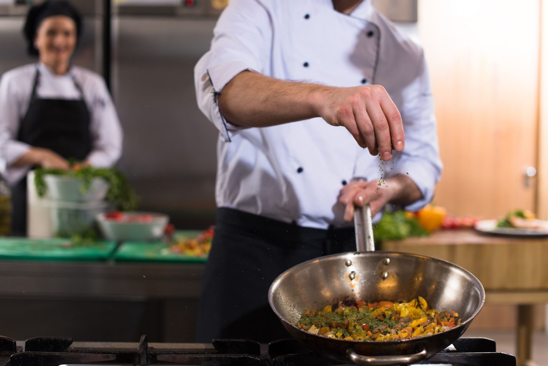 Un chef con uniforme blanco espolvoreando especias en una sartén sobre una estufa, con otro chef al fondo.