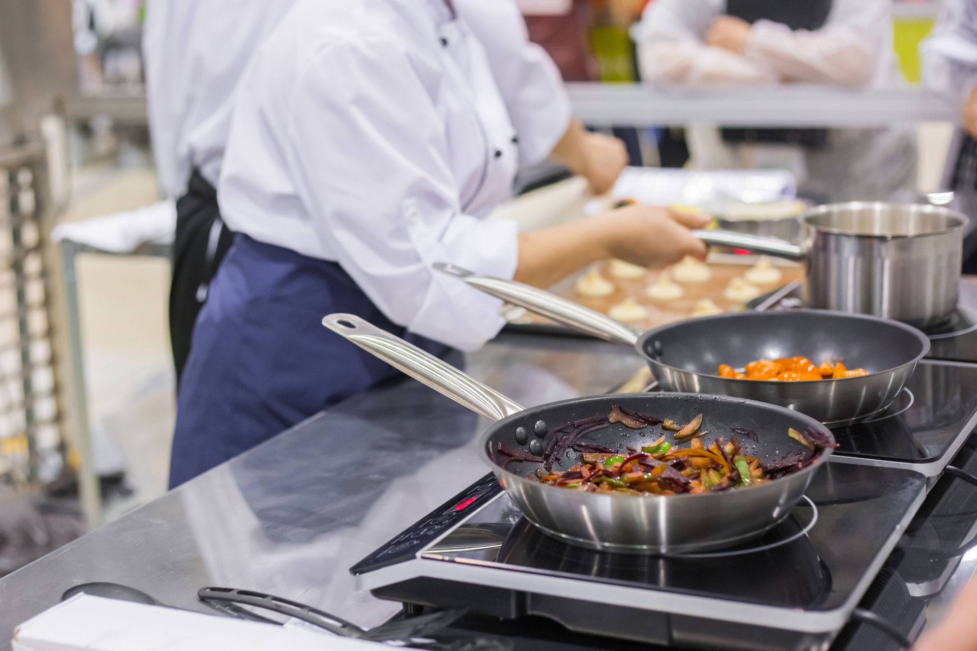 Chefs con batas blancas y delantales cocinando en una cocina comercial; sartenes con comida sobre una estufa, alguien sosteniendo una bandeja.