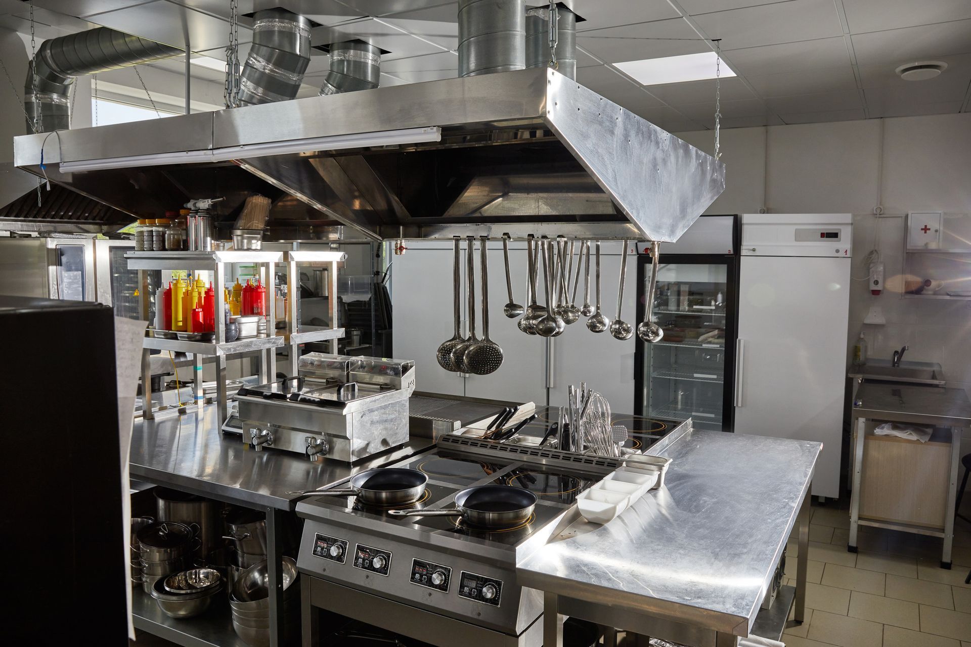 Una cocina comercial con electrodomésticos de acero inoxidable, incluyendo estufas, hornos y campanas extractoras. Ollas y cucharones cuelgan sobre la zona de cocción.