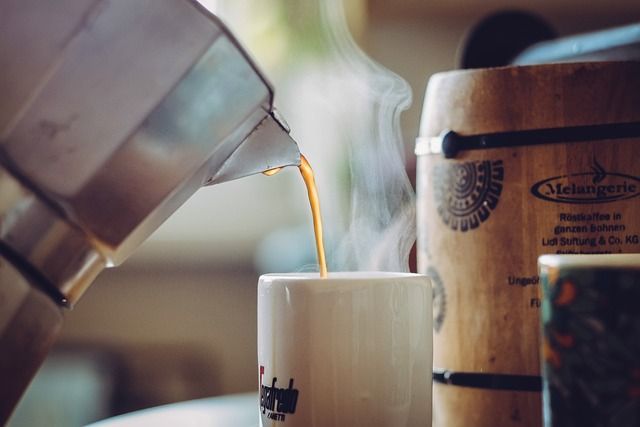 Café vertido desde una cafetera expreso sobre una taza blanca, con vapor elevándose junto a un recipiente de madera y parte de una taza decorada.