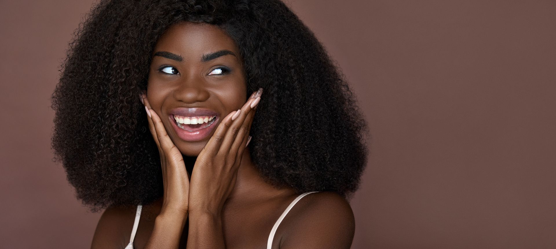 Femme à la peau foncée, souriante et touchant ses joues, sur fond marron.