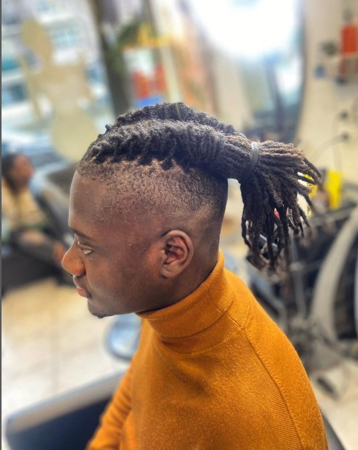 Homme aux dreadlocks foncées coiffées en queue de cheval, coupe dégradée undercut, portant un col roulé orange.