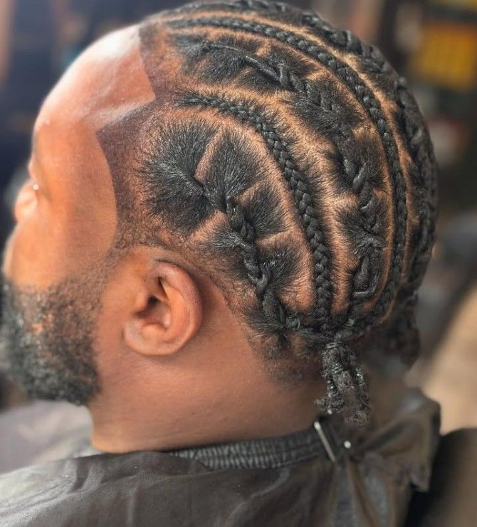 Homme avec une coiffure tressée, un motif rasé sur le côté de la tête, un bouc et portant une cape de barbier noire.