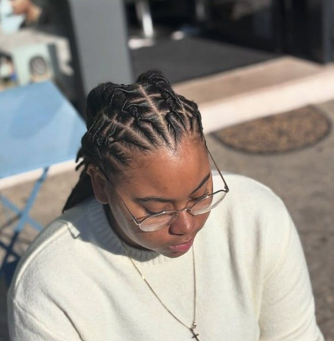 Femme portant des lunettes, un pull crème et une coiffure tressée. En extérieur.