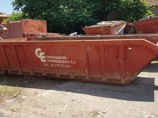 Contenedor de obra grande y rojo en una calle soleada, con árboles al fondo