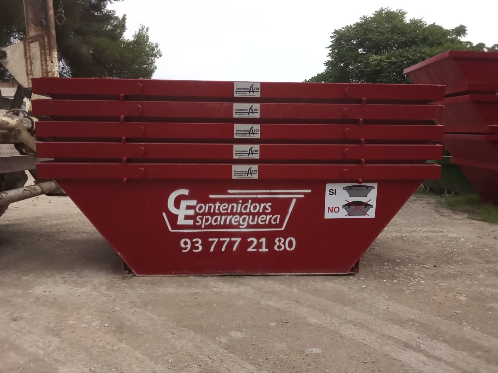 Contenedor de obra rojo con logotipo blanco y número de teléfono en un terreno de tierra
