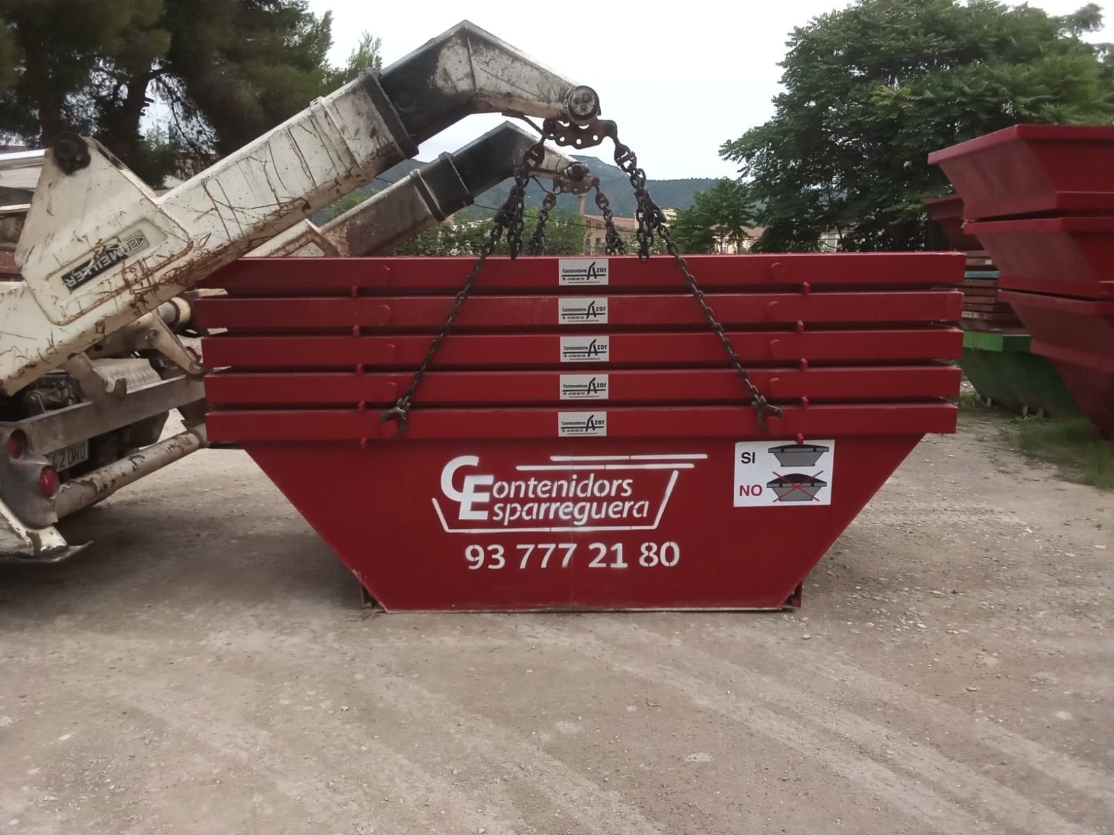 Un contenedor rojo de obra que está siendo llenado por un camión volquete en un camino de tierra