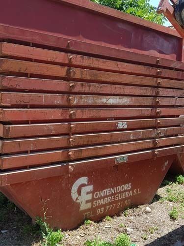 Contenedor rojo de obra con listones laterales de madera y el logotipo de una empresa española sobre un terreno de grava