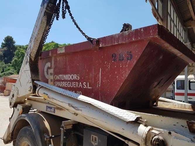 Contenedor rojo de obra sobre un camión de plataforma en una zona de trabajo, con el brazo de una grúa levantándolo.
