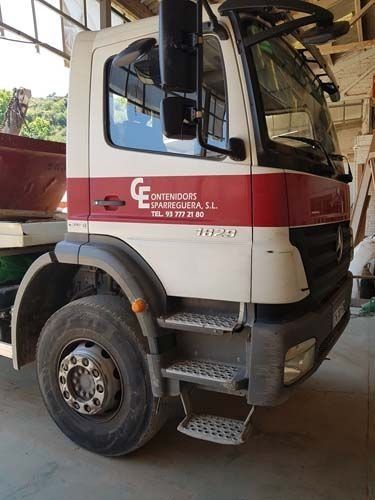Camión rojo y blanco con cabina sobre el motor, estacionado en interiores, visto de lado y con el logotipo de la empresa en la puerta.