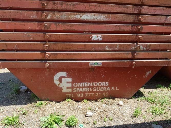 Contenedor de obra metálico rojo con el logotipo y el nombre de la empresa en blanco en un lateral, al aire libre sobre el césped.