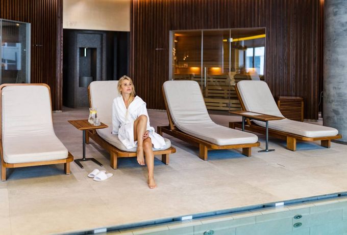 Mujer con bata blanca sentada junto a una piscina cubierta, al lado de tumbonas vacías.