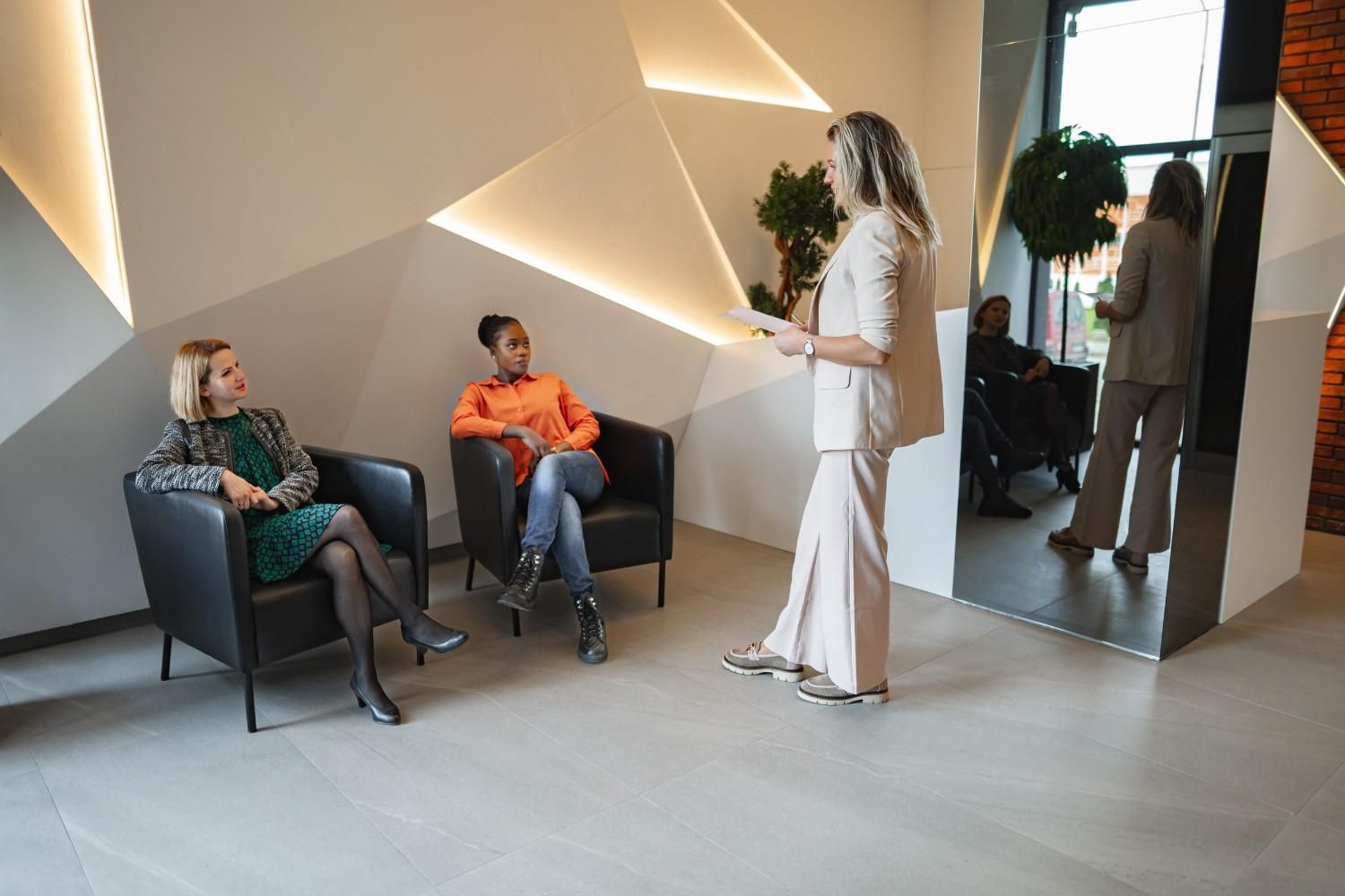 Tres personas en un vestíbulo moderno, dos sentadas y una de pie, con iluminación geométrica en la pared.
