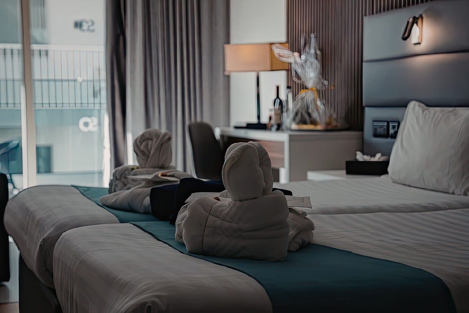 Habitación de hotel con dos cisnes hechos con toallas sobre una cama impecablemente hecha, lámparas y cortinas al fondo.