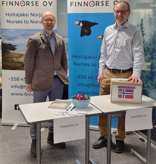 Kaksi rekrytoijaa seisoo kahden "Finnorse Oy" -bannereiden kanssa varustetun pöydän takana sisätiloissa. Toinen pitelee Norjan lipulla varustettua laatikkoa.