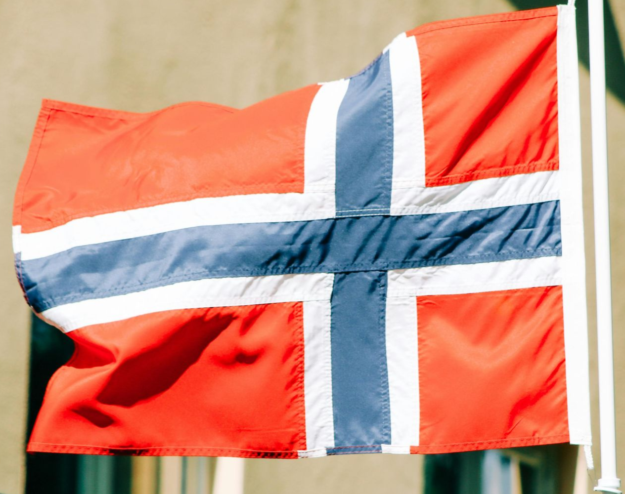 Norwegian flag waving in the breeze; red with blue cross edged in white, on a flagpole outdoors.