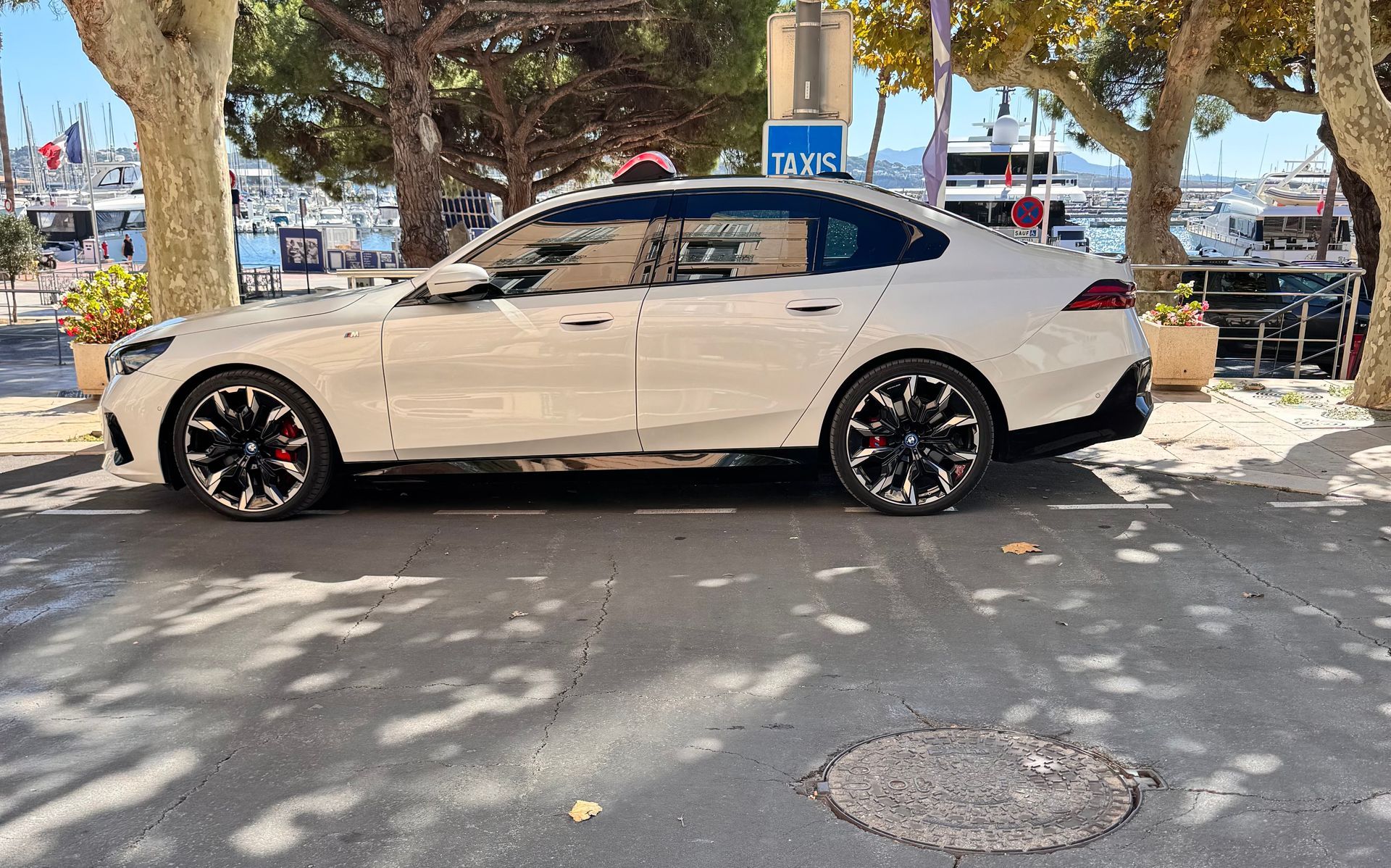 Une berline blanche, aux jantes noires, est garée dans une rue de la ville. On aperçoit des arbres et des bateaux à l'arrière-plan.