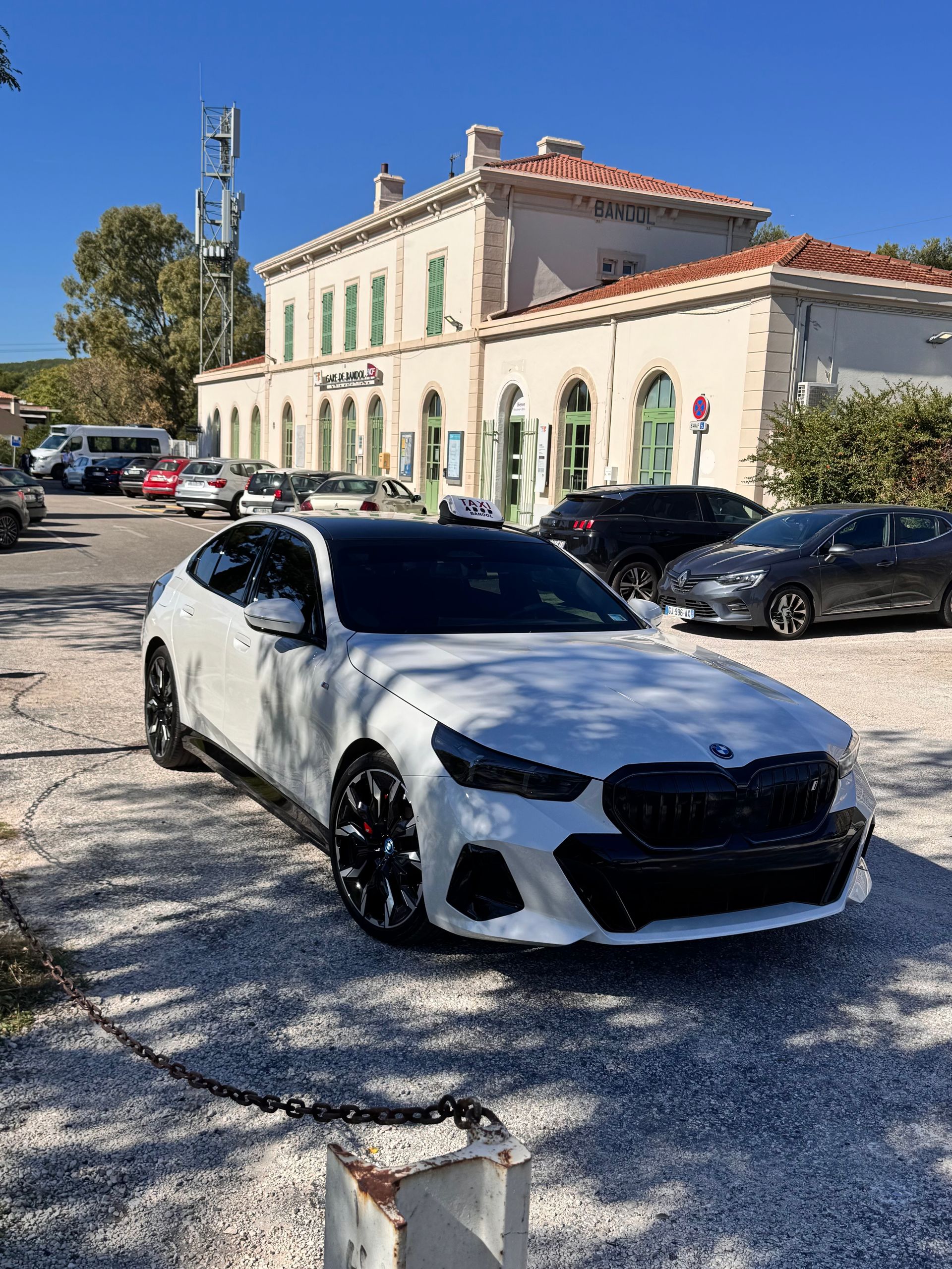 Une BMW blanche est garée devant un bâtiment clair, avec des voitures et un ciel dégagé en arrière-plan.