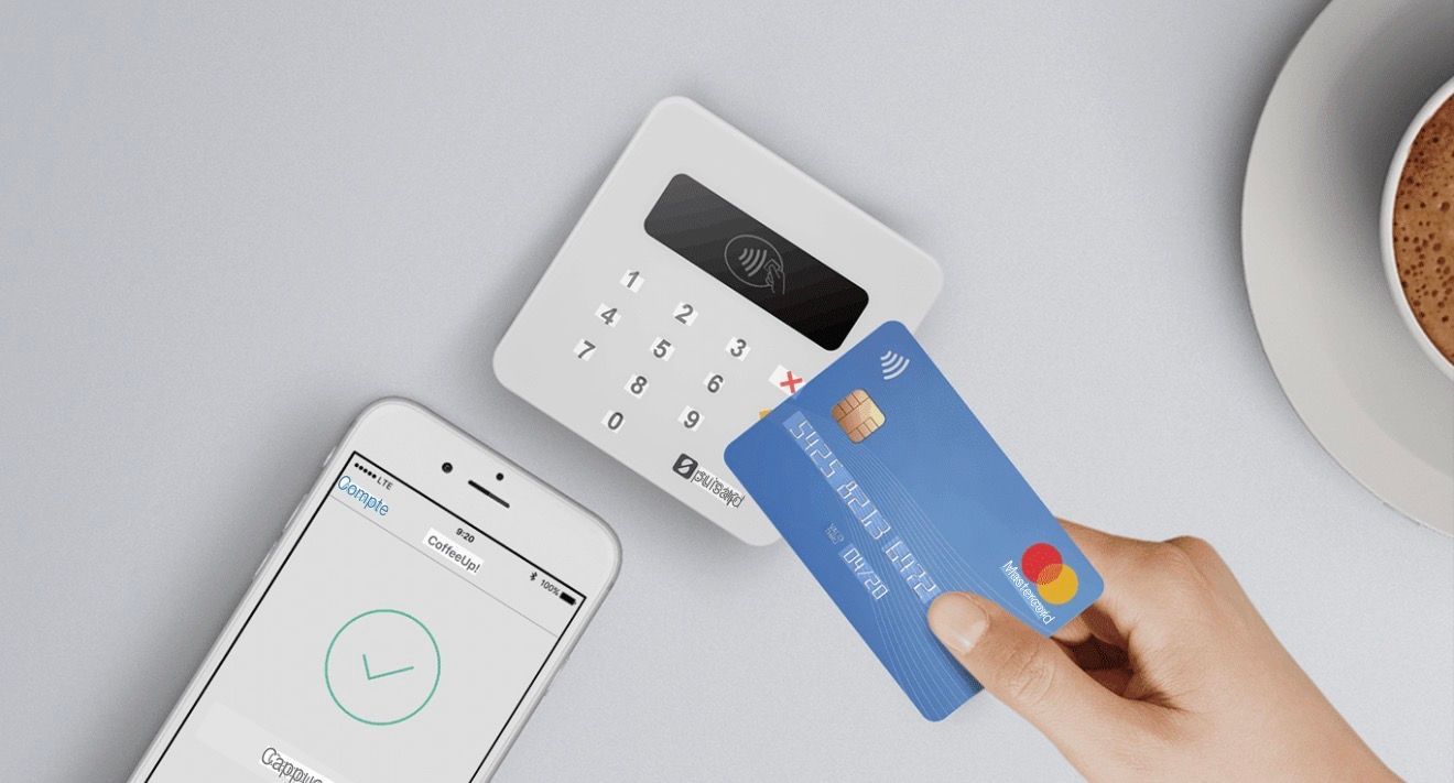 Une main tenant une carte de crédit bleue près d'un terminal de paiement, d'un téléphone et d'une tasse.