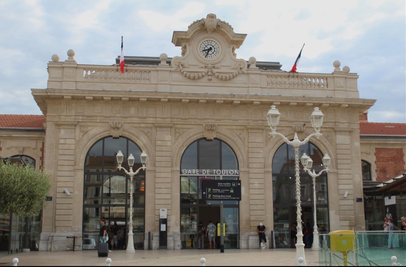 Extérieur d'une gare en pierre claire avec entrées voûtées et horloge. Drapeaux français au sommet.