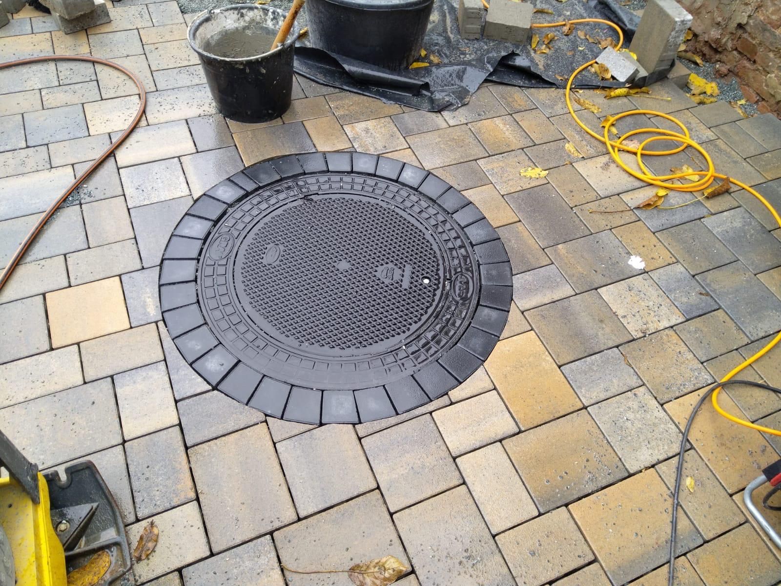 Runder Kanaldeckel, umgeben von Pflastersteinen. Bauwerkzeuge und -materialien sind sichtbar.
