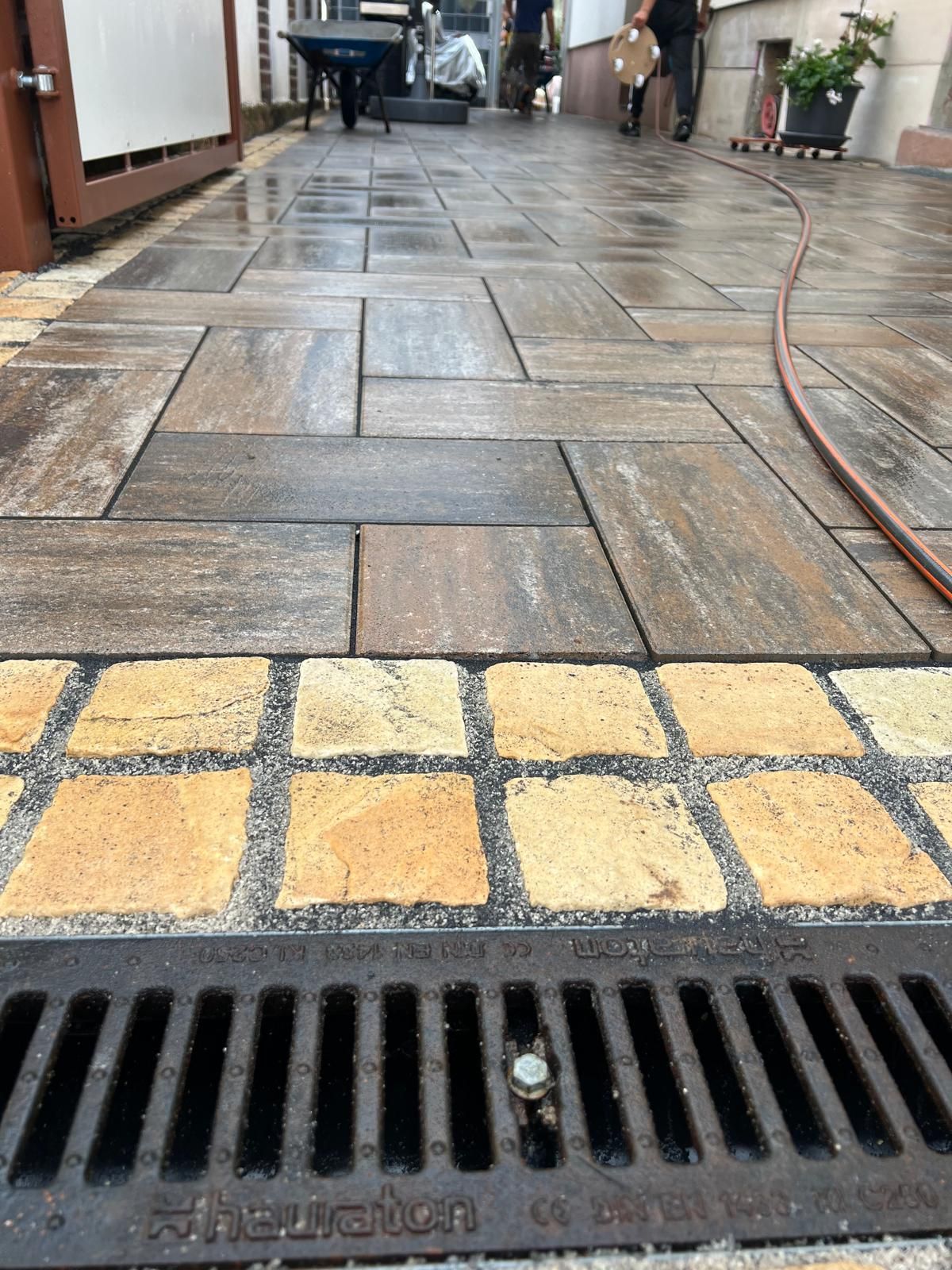 Gepflasterter Gehweg mit einem Abflussgitter am Ende. Arbeiter im Hintergrund, nasse Steinplatten und ein Rand aus gelben Ziegelsteinen.