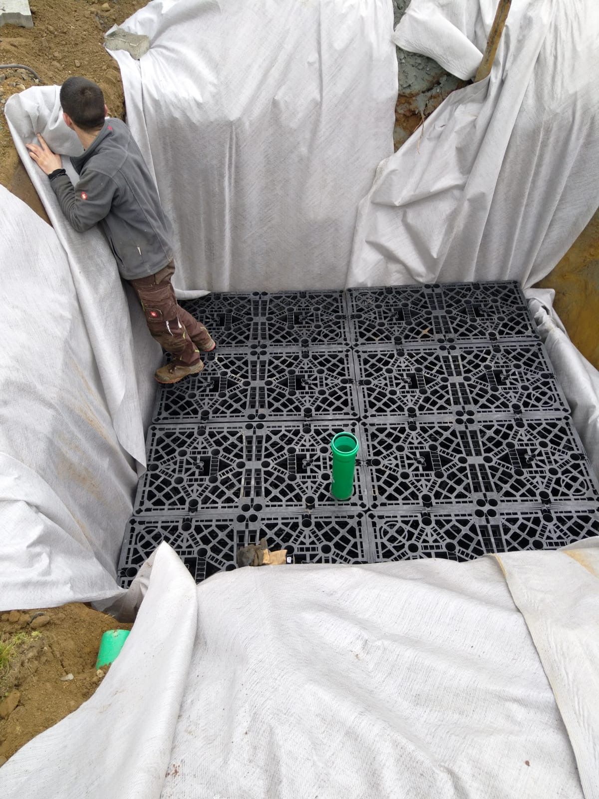 Ein Mann verlegt ein mit weißem Stoff abgedecktes Kunststoff-Drainagegitter in einen Graben.
