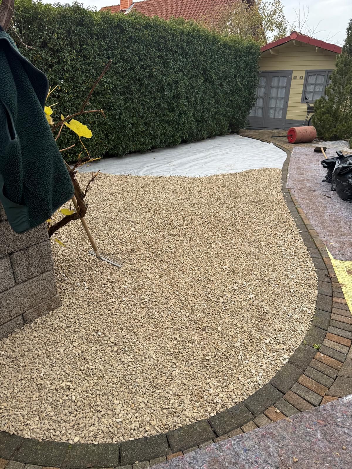 Ein mit Kies bedecktes Gartenbeet mit geschwungener Ziegelsteinumrandung, vor einer grünen Hecke und einem kleinen Schuppen.